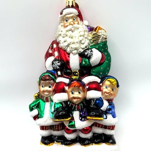May include: A Christmas ornament featuring Santa Claus with a white beard and red suit, holding a green sack of gifts. Below Santa are four elves in green, red, and blue outfits. The ornament is topped with a gold star.