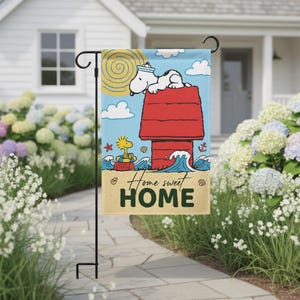 May include: A decorative garden flag featuring Snoopy in a sailor hat atop his red doghouse, with Woodstock in a boat. The flag has a blue sky background with the words "Home sweet HOME" in a sand-colored font.