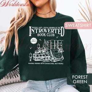 May include: Forest green sweatshirt with white graphic text: "PROUDLY ANTI-SOCIAL MEMBER OF THE INTROVERTED BOOK CLUB" and "MAKING FRIENDS WITH CHARACTERS, NOT PEOPLE". The design includes books, candles, and floral elements. The word "SWEATSHIRT" is in the upper right corner.