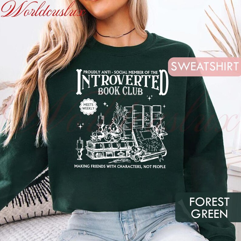 May include: Forest green sweatshirt with white graphic text: "PROUDLY ANTI-SOCIAL MEMBER OF THE INTROVERTED BOOK CLUB" and "MAKING FRIENDS WITH CHARACTERS, NOT PEOPLE". The design includes books, candles, and floral elements. The word "SWEATSHIRT" is in the upper right corner.