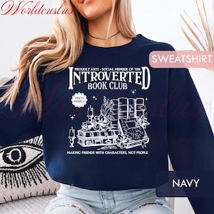 May include: Navy blue sweatshirt with white graphic text: "INTROVERTED BOOK CLUB" and "MAKING FRIENDS WITH CHARACTERS, NOT PEOPLE." The design includes illustrations of books, candles, and floral elements. The word "SWEATSHIRT" is in a pink box.