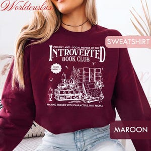 May include: Maroon sweatshirt with a white graphic that reads "Proudly Anti-Social Member of the Introverted Book Club." The design includes illustrations of books, flowers, and candles. The text "Making Friends With Characters, Not People" is also included.