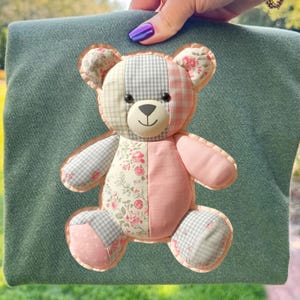 Può includere: Applicazione di orsetto patchwork dai colori pastello, tra cui rosa, grigio e motivi floreali, su sfondo verde. L'orso ha un volto sorridente ed è realizzato con vari tessuti.
