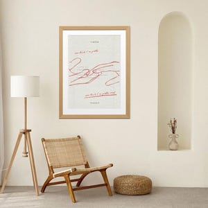 May include: A framed art print with a light wood frame. The artwork features red line drawings of hands and text that reads "you think I'm pretty" and "you think I'm pretty weak". The print hangs on a cream-coloured wall.