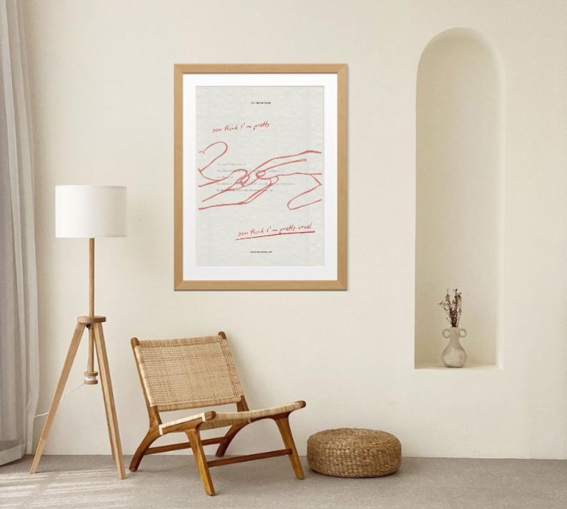 May include: A framed art print with a light wood frame. The artwork features red line drawings of hands and text that reads "you think I'm pretty" and "you think I'm pretty weak". The print hangs on a cream-coloured wall.
