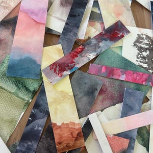 May include: A collection of watercolor bookmarks in various colors and designs. The bookmarks feature abstract landscape and color-field paintings in shades of blue, pink, red, green, and brown. The bookmarks are arranged in a scattered pattern.