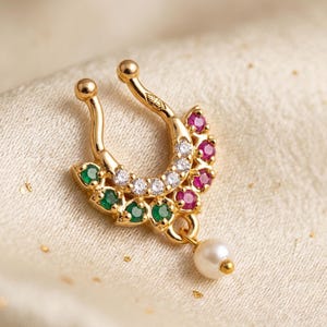 May include: A gold-toned septum ring featuring a horseshoe shape adorned with a row of clear stones, three green stones on the left, three pink stones on the right, and a single dangling white pearl-like bead at the bottom center.