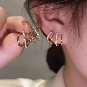Può includere: Un ear cuff color oro dal design multistrato composto da tre fasce curve distinte. Ogni fascia è incastonata con piccole pietre chiare e scintillanti che catturano la luce. Il gioiello è progettato per essere indossato sul cartilagine.