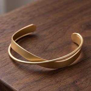 Könnte beinhalten: Ein goldfarbenes Armband in Manschettenform mit matter Oberfläche. Das Design besteht aus zwei überlappenden Bändern, die sich vorne kreuzen und eine offene, kreisförmige Form mit abgerundeten, glatten Enden an beiden Seiten bilden.
