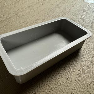 May include: A rectangular, gray baking pan with a textured rim. The pan is empty and sits on a wooden surface. The pan's interior is smooth and the corners are slightly rounded.