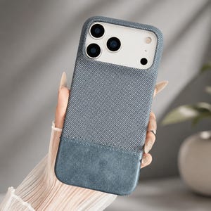May include: A smartphone case featuring a textured blue fabric upper section and a smooth, solid blue leather-like lower panel. The case includes a large rectangular cutout at the top to accommodate the camera lenses and flash of the mobile device.