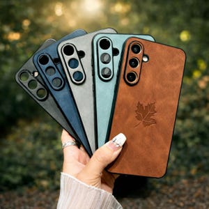 Puede incluir: Una colección de cinco fundas para teléfono en varios colores: negro, azul marino, gris, verde azulado y marrón. La funda marrón presenta un diseño de hoja. Cada funda tiene un recorte para la cámara.