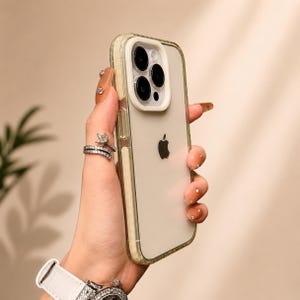 Puede incluir: Una funda protectora transparente para un iPhone de Apple con un módulo de cámara blanco que alberga tres lentes. La funda cuenta con un panel trasero transparente y un marco de color claro, mostrando el logotipo negro de Apple en el centro.