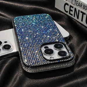 Puede incluir: Una funda de teléfono adornada con pedrería azul y plateada en degradado. La funda tiene un borde negro y un marco plateado alrededor de las lentes de la cámara. La funda del teléfono está sobre una superficie de satén negro.