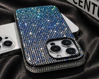 May include: A bedazzled phone case with a gradient of blue and silver rhinestones. The case has a black border and a silver frame around the camera lenses. The phone case is on a black satin surface.