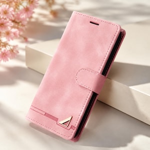 Puede incluir: Una funda de teléfono tipo cartera de color rosa claro con un acabado mate suave, que cuenta con una solapa de cierre magnético y un pequeño emblema metálico triangular de tono dorado en la esquina inferior. La funda tiene costuras visibles.