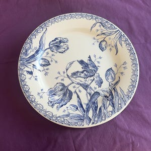 May include: A white ceramic plate with a blue floral design featuring tulips and foliage. The plate has a detailed border and the floral pattern is repeated around the edge. The design is in a classic, vintage style.