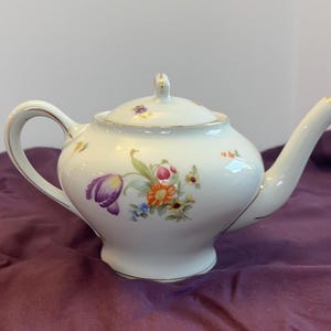 May include: White porcelain teapot with a lid, handle, and spout. The teapot is decorated with a floral design featuring purple, orange, and yellow flowers. Gold trim accents the edges of the teapot and handle.