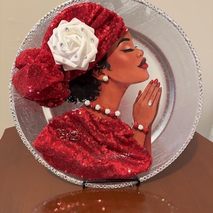 Peut inclure: Une assiette de présentation décorative argentée ornée d'une illustration de profil d'une personne à la peau foncée, les yeux fermés en prière, portant un foulard à sequins rouges avec une rose blanche, un haut à sequins rouges et un collier.