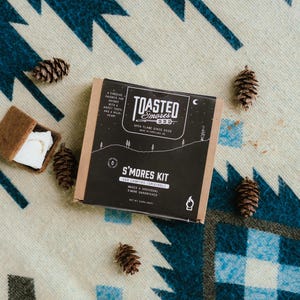 Puede incluir: Una caja negra y beige de kit S'mores con el texto "Toasted S'mores" y "S'mores Kit", rodeada de piñas y un s'more. El kit está sobre una manta estampada con acentos azules, blancos y rojos.