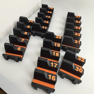 Op de afbeelding: Zwarte en oranje genummerde blokken, waarschijnlijk voor een spel of organisatiesysteem. Elk blok heeft een zwarte behuizing met oranje accenten en een oranje nummer. De nummers variëren van 6 tot 32.