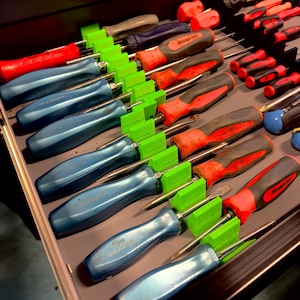 May include: A selection of screwdrivers with red, blue, and black handles, arranged in a drawer. The screwdrivers are secured by green holders. Some screwdrivers feature the "Snap-on" logo.