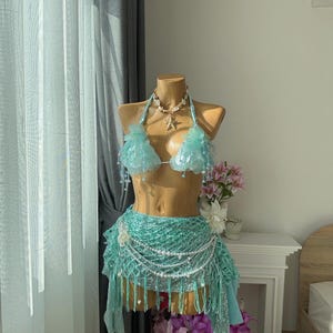 May include: A mermaid-themed costume on a mannequin. The outfit features a light blue seashell bikini top, a matching skirt with fringe and pearl details, and a seashell necklace. The costume is designed with a fantasy theme.