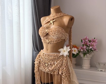 Op de afbeelding: Een beige tweedelig danskostuum met gouden versieringen, weergegeven op een mannequin. De set bevat een bh-top met zeesteraccenten, een rok met hoge taille en een gedrapeerde sjaal. Een witte bloem siert de taille.