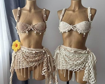 May include: Two handmade crochet bikini sets displayed on mannequins. One is beige, the other is off-white. Both feature pearl accents and matching skirts with fringe and draping. A yellow flower adorns one set.