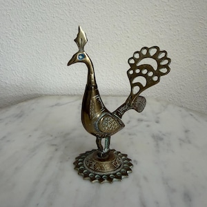 May include: A brass peacock figurine featuring an intricate openwork tail, a crest on its head, and a small blue inlay for the eye. The bird stands on a circular, scalloped base with engraved patterns, showcasing a metallic gold finish.