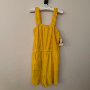 May include: A bright yellow sleeveless romper made of a textured terry cloth fabric. The garment features wide shoulder straps with button details, an elasticated waist, and a single patch pocket on the front right side. A white clothing tag is attached.