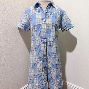 May include: A short-sleeved button-down dress featuring a patchwork design composed of various blue, white, and pale yellow fabric squares. The garment includes a classic collar, a full-length front button placket, and a knee-length hemline.