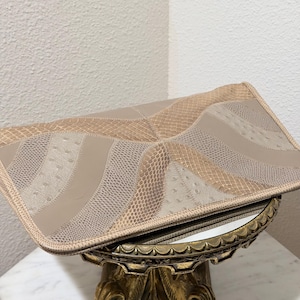 May include: A rectangular clutch bag featuring a geometric patchwork design in shades of beige and tan. The surface alternates between smooth panels and textured sections with a reptilian scale pattern and small raised dots throughout the exterior.