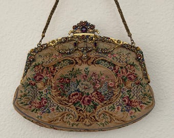 May include: A beige tapestry handbag featuring a floral needlepoint design with pink, red, and blue flowers. The gold-tone metal frame is embellished with small colorful stones and a central purple oval gem, complemented by a thin gold chain strap.