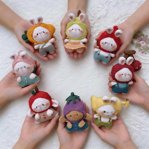 Peut inclure: Plusieurs poupées lapins crochetées à la main, chacune vêtue d'une tenue unique sur le thème des fruits ou des légumes. Les poupées ont des corps blancs, des joues roses et sont tenues dans différentes mains. Les tenues comprennent une carotte, une poire, une pomme et une aubergine.
