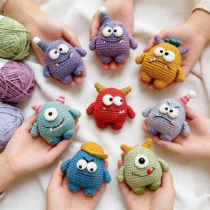 Op de afbeelding: Een verzameling handgemaakte gehaakte monstertjes in verschillende kleuren, waaronder paars, rood, geel, blauw en groen. Elk monster heeft een unieke uitdrukking en is versierd met kleine hoedjes of hoorns. De speeltjes worden in handen gehouden, wat hun grootte en details laat zien.