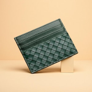 May include: A dark green card holder featuring a woven leather texture on the lower half and two horizontal card slots on the upper portion. The accessory has visible tonal stitching along the edges and a rectangular shape with a slim profile.