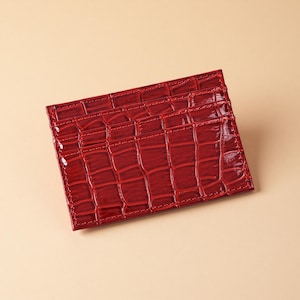 Può includere: Un porta carte rosso con una texture lucida effetto pelle di coccodrillo. Il portafoglio rettangolare ha più fessure per carte ed è realizzato in un materiale lucido e resistente. I bordi sono cuciti con filo rosso.