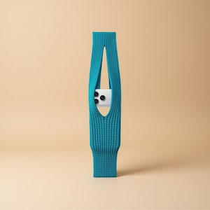 Può includere: Un porta telefono in maglia color teal con un design verticale. Il supporto ha una forma lunga e rettangolare con un ritaglio al centro per contenere un telefono. Il telefono è bianco e ha tre obiettivi della fotocamera. Lo sfondo è beige.