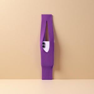 Può includere: Una vivace custodia per telefono in maglia viola dal design unico. La custodia ha una forma lunga e rettangolare con un'apertura centrale per contenere un telefono. Il telefono è bianco con due lenti nere.