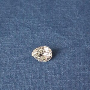 May include: A pear-shaped, clear gemstone with multiple facets, resting on a textured blue surface. The gemstone is cut to maximize light reflection, creating a sparkling effect. The gem is a light color.