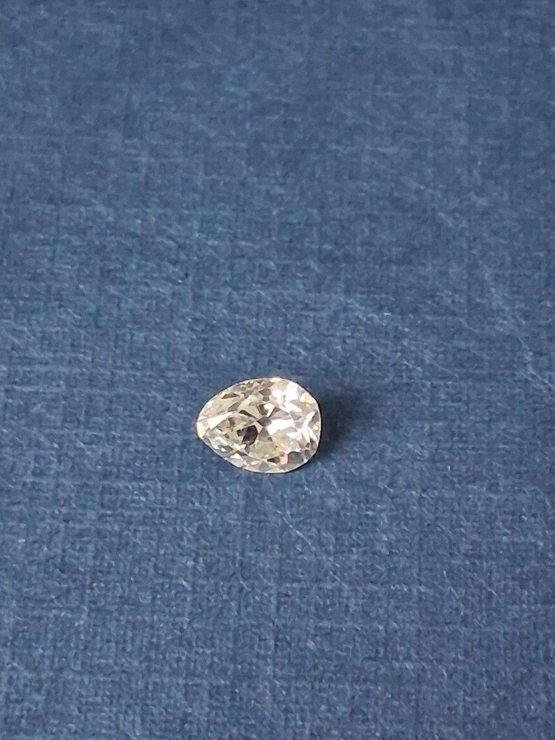 May include: A pear-shaped, clear gemstone with multiple facets, resting on a textured blue surface. The gemstone is cut to maximize light reflection, creating a sparkling effect. The gem is a light color.