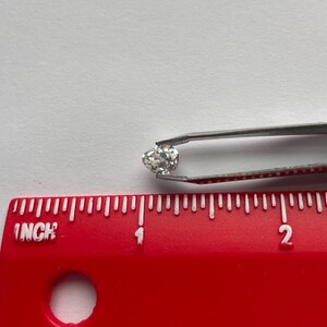 May include: A small, round, clear diamond held by silver tweezers. The diamond is positioned near a red ruler with inch markings. The ruler is placed on a white surface.