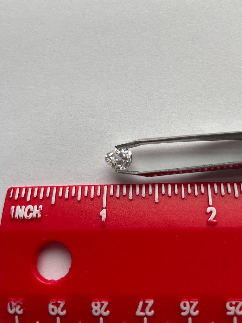May include: A small, round, clear diamond held by silver tweezers. The diamond is positioned near a red ruler with inch markings. The ruler is placed on a white surface.
