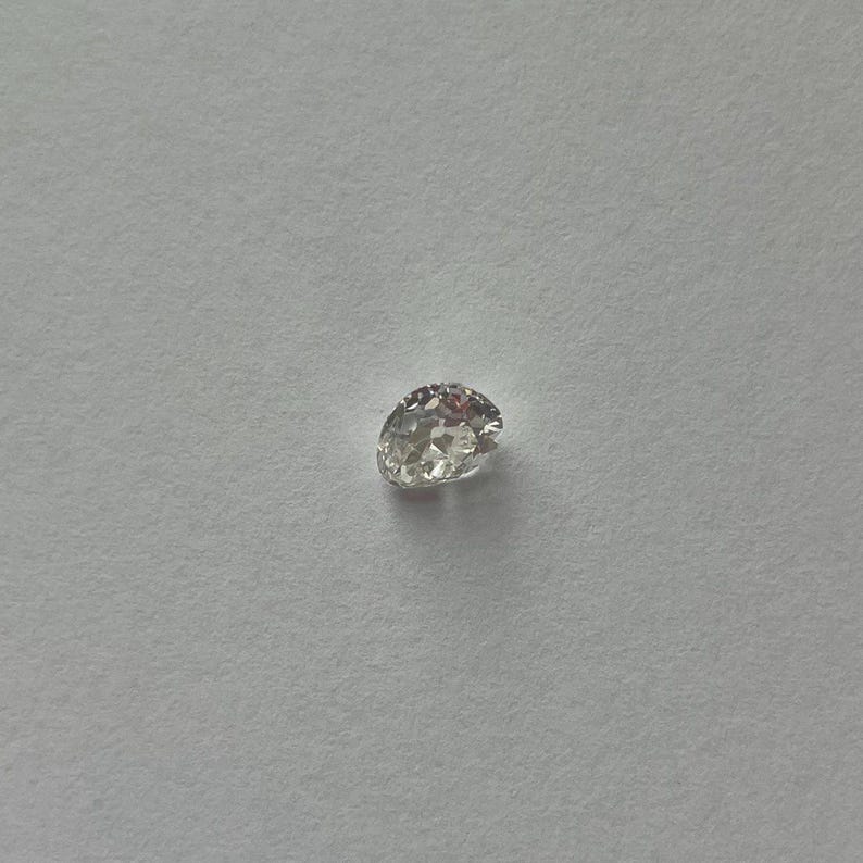 May include: A single, clear, oval-shaped diamond with multiple facets. The gemstone is set against a plain, light gray background, showcasing its brilliance and clarity. The diamond appears to be a loose stone, ready for setting.