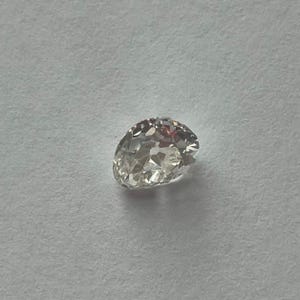 May include: A clear, oval-shaped diamond with multiple facets, reflecting light. The gemstone is set against a plain, light-colored background, showcasing its brilliance and clarity. This diamond is suitable for jewelry.