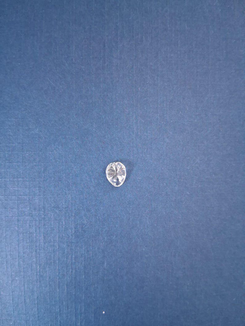 May include: A clear, oval-shaped gemstone with multiple facets, reflecting light. The gem is set against a textured, dark blue background, creating a contrast that highlights its brilliance. This gemstone could be used in jewelry.
