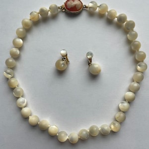 May include: A vintage jewelry set featuring a cream-colored beaded necklace and matching clip-on earrings. The necklace has a gold clasp with a cameo. The earrings have a pearl-like appearance.