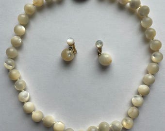 May include: A vintage jewelry set featuring a cream-colored beaded necklace and matching clip-on earrings. The necklace has a gold clasp with a cameo. The earrings have a pearl-like appearance.