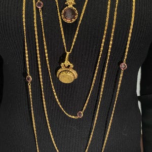 May include: Several gold necklaces of varying lengths, some with small, round, purple gemstones. One necklace features a pendant with a round, purple gemstone. Another has a coin-shaped pendant. The necklaces are layered against a black background.